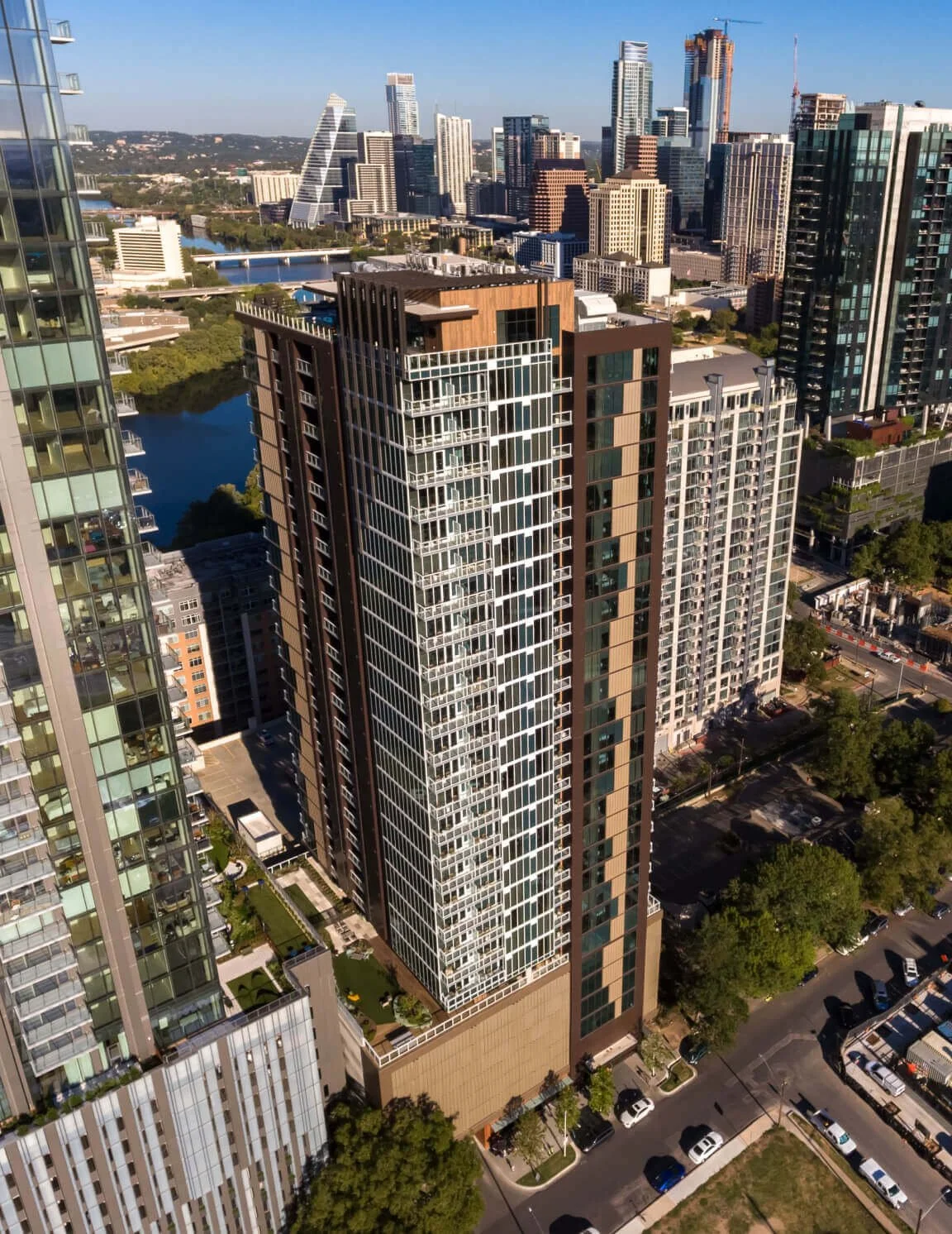 Aerial view of a modern high-rise building in an urban city with other tall buildings, a river, and a park in the background.