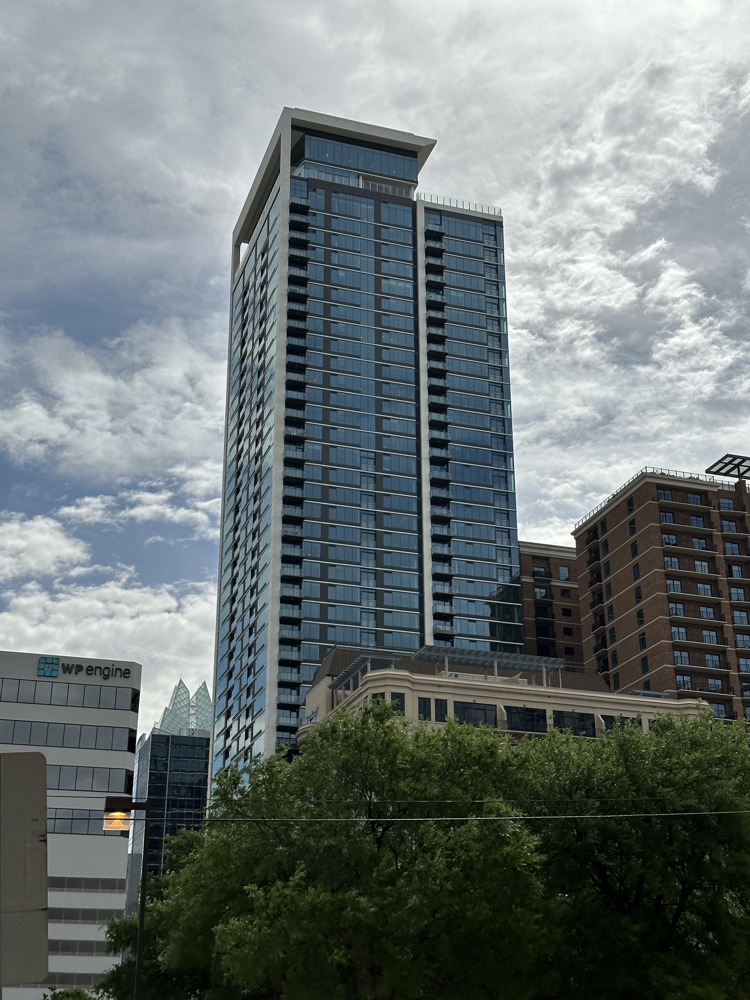 Tall modern skyscraper with glass windows and a unique overhanging roof, surrounded by other high-rise buildings and trees, under a cloudy sky.