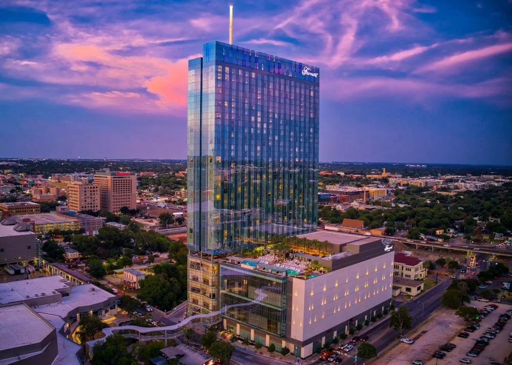 Nighttime aerial view of a tall, glass-encased hotel building with a pool and rooftop area, surrounded by cityscape and a colorful sunset sky.