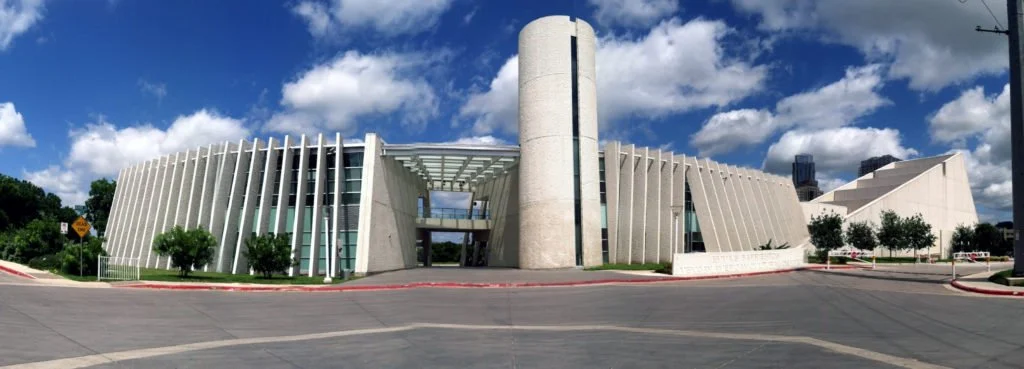 A modern, architecturally unique building with curved white exterior walls and a tall cylindrical tower in the center, set against a bright blue sky with scattered clouds.