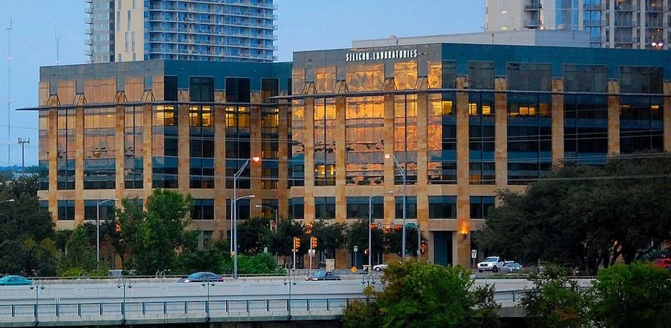 Modern office building with reflective glass windows and beige stone facade, with trees and cars in the foreground during sunset.
