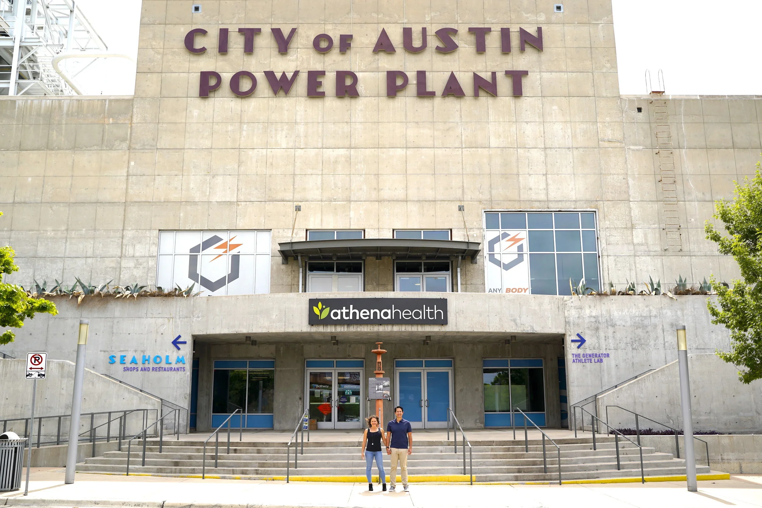 Two people standing in front of the City of Austin Power Plant building with signs for Athena Health, Seaholm Shops and Restaurants, and The Generator Athlete Lab.