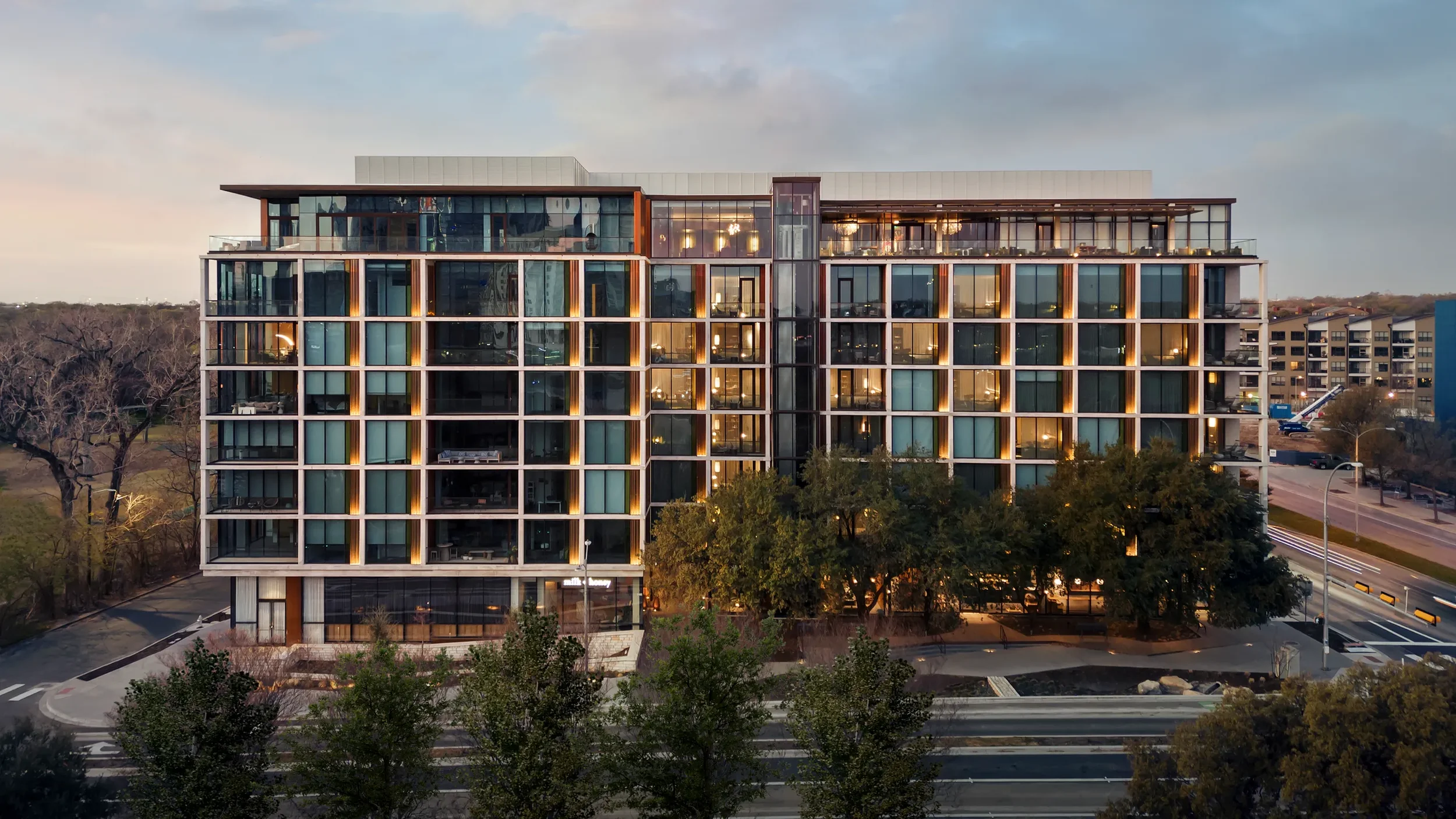 Modern multi-story apartment building with glass facade, lit windows, trees in front, situated by a street with moving cars, in early evening.