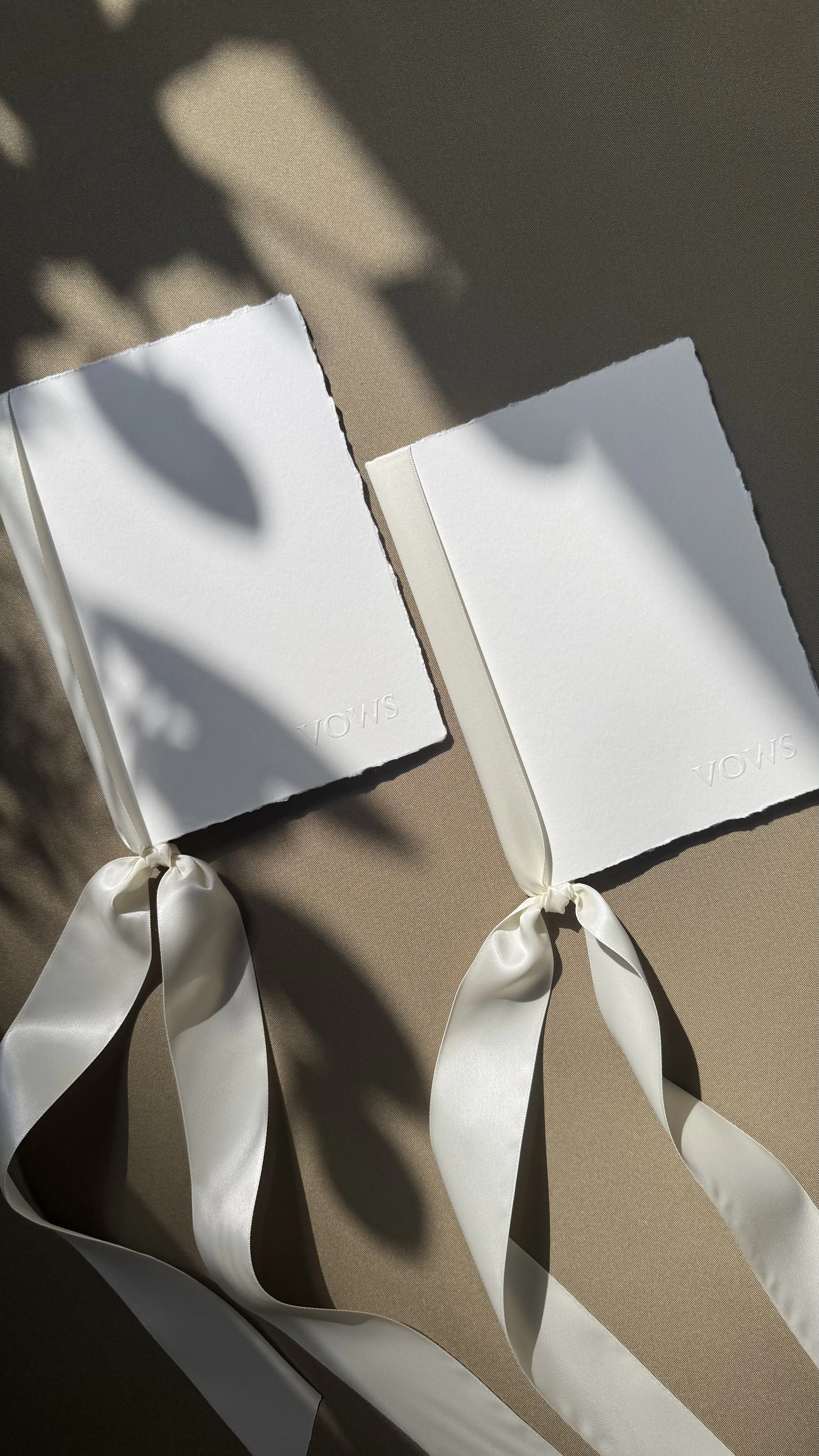 Two white notebooks with embossed text 'S.MODA' and satin ribbons, placed on a beige surface with shadow of leaves.