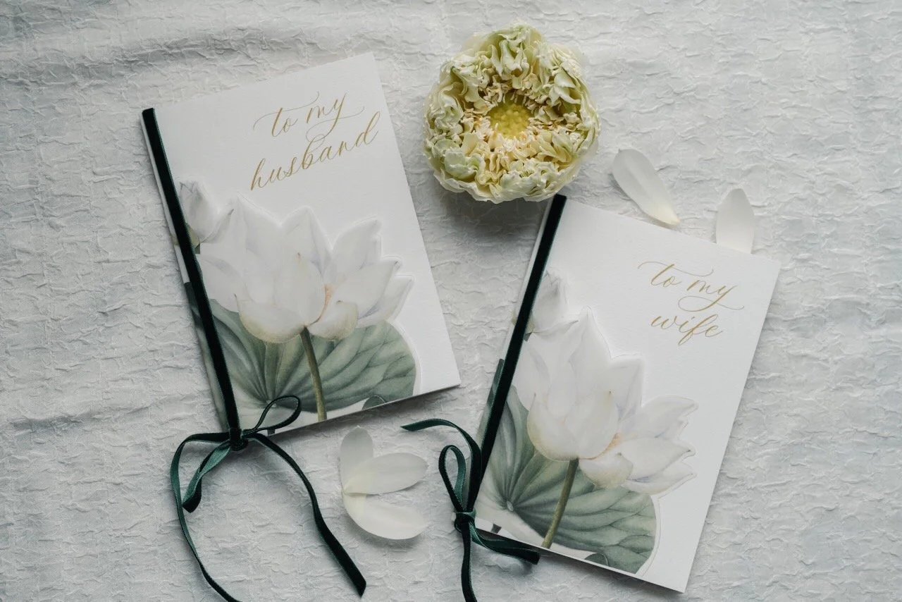 Two greeting cards with floral designs, one says "to my husband" and the other "to my wife," accompanied by white flower petals and a white flower in a small pot on a textured white surface.