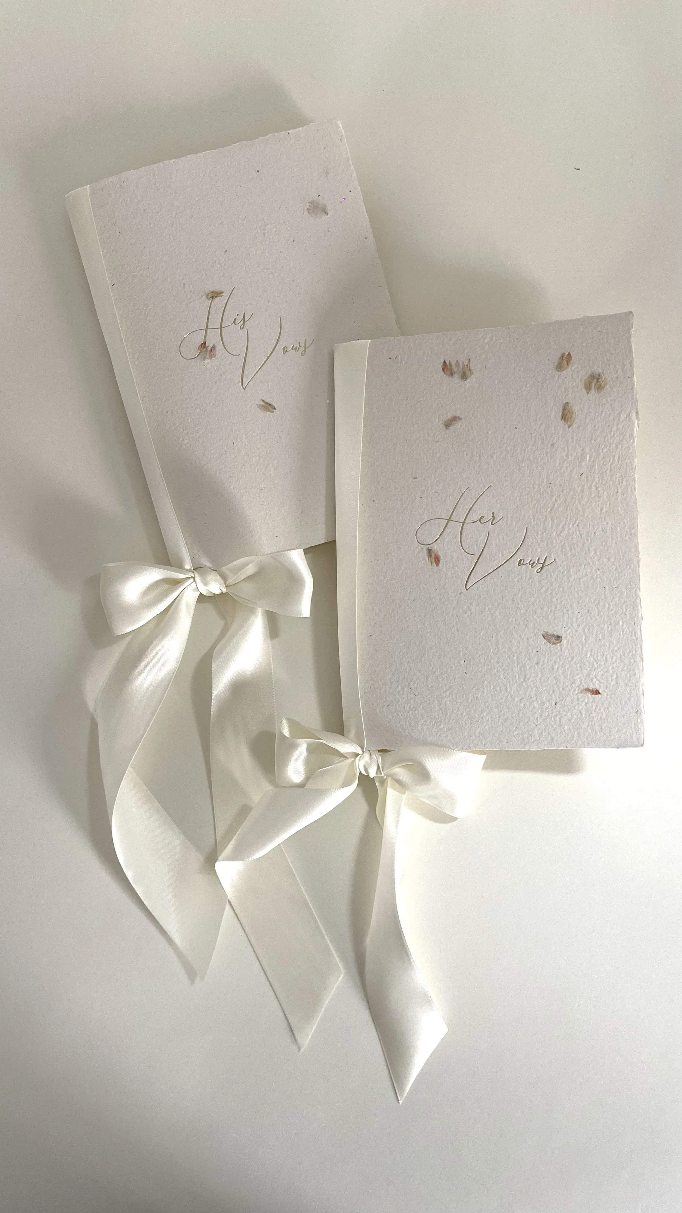 Two white textured greeting cards, one labeled 'His Love' and the other 'Her Love' in gold script, each tied with a white satin ribbon bow, with small dried flower petals scattered on top.