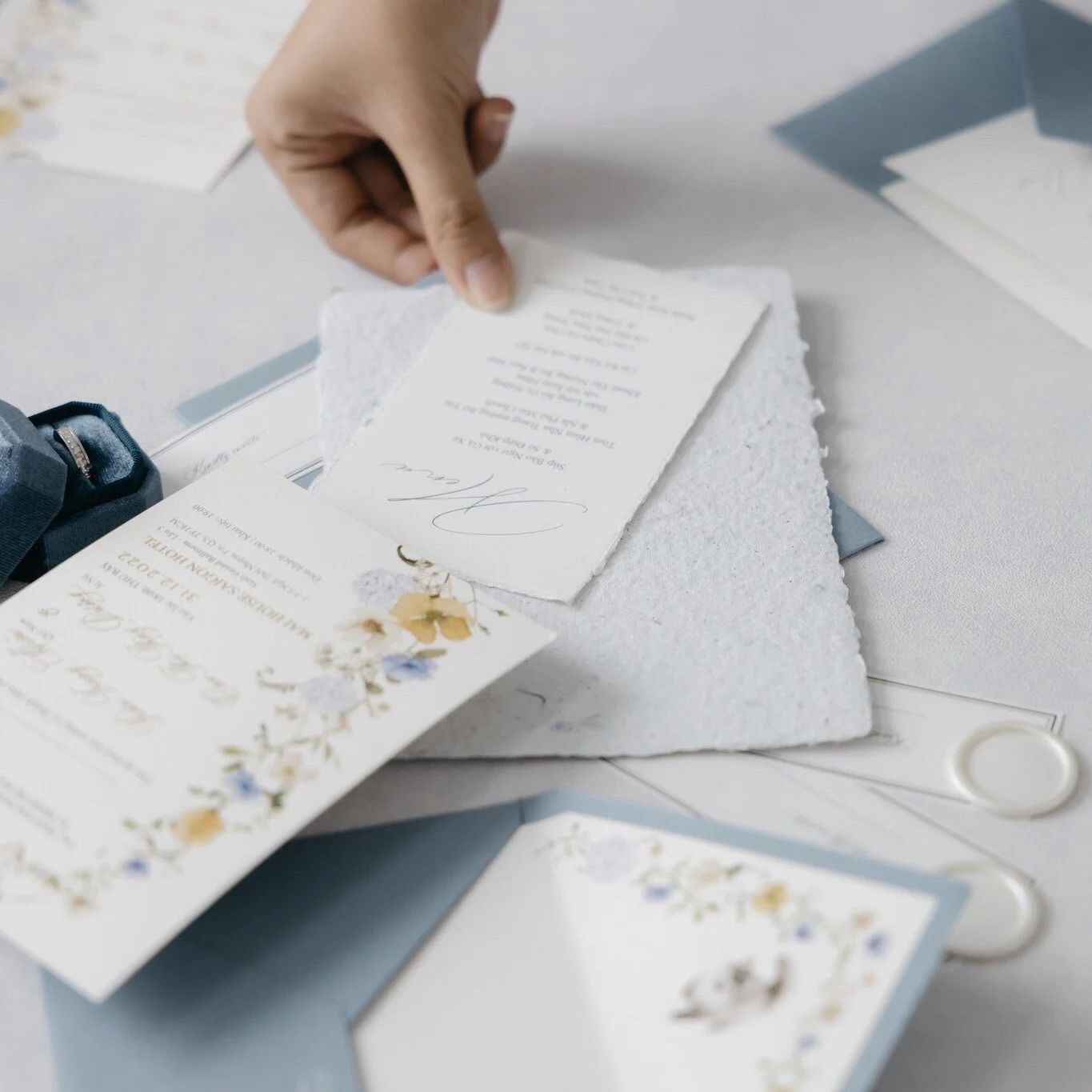 Hand placing a white textured envelope on a table with invitations and cards, decorated with floral designs, for a wedding or event.