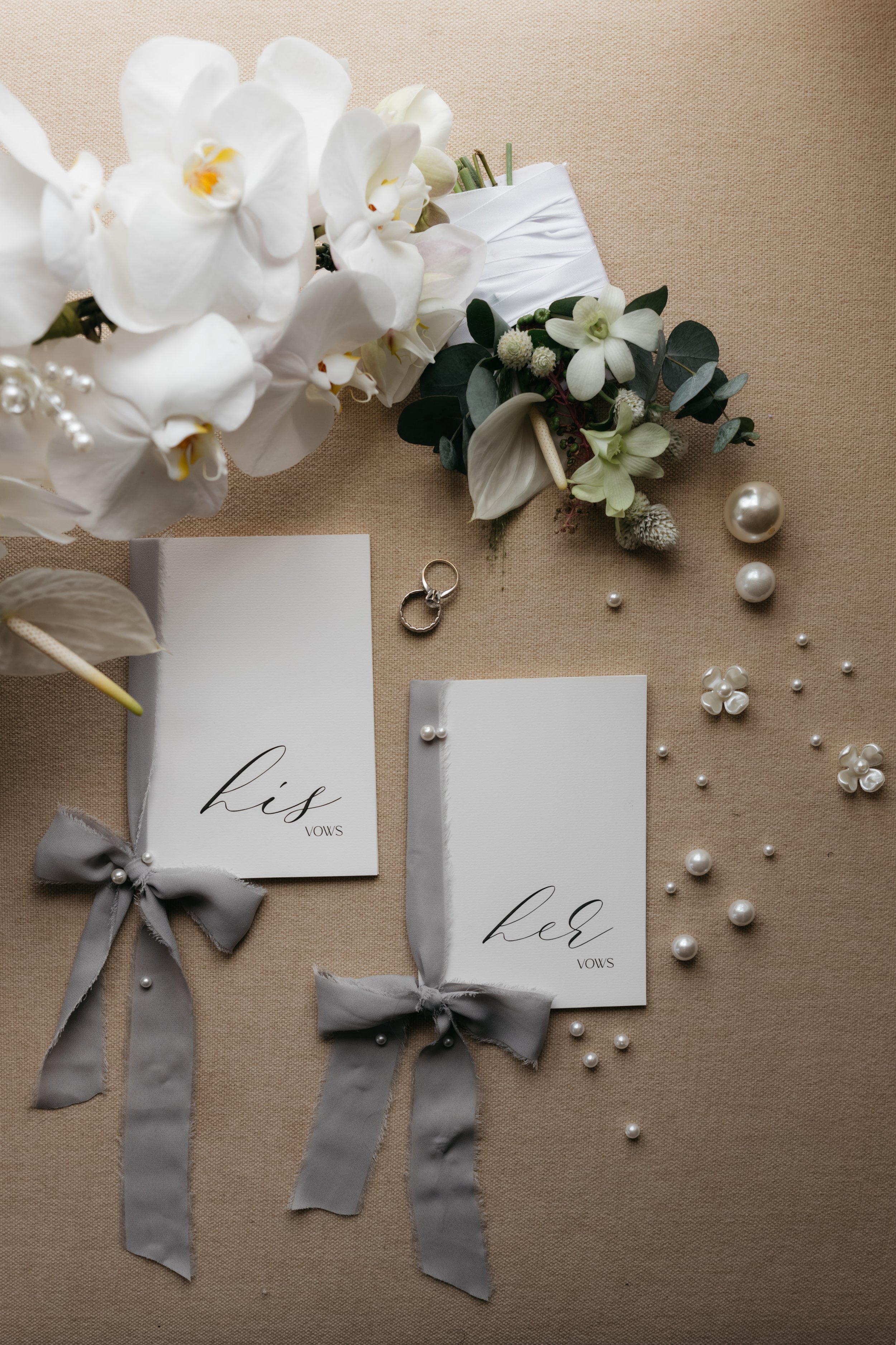 Wedding vow cards for the bride and groom with floral decorations, pearls, and rings on a beige fabric background.