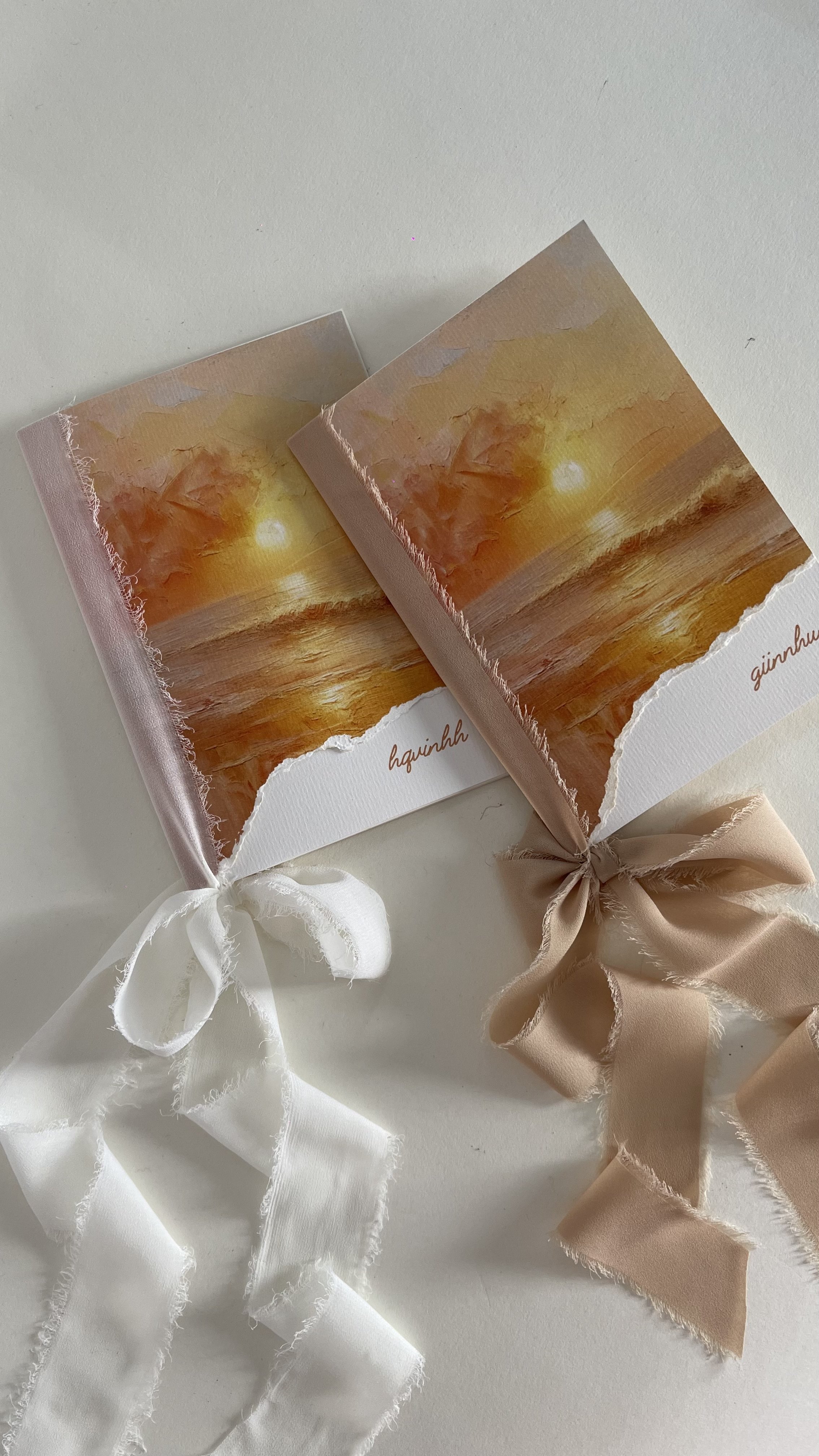Two notebooks with sunset artwork on the covers, tied with white and beige satin ribbons, placed on a white surface.