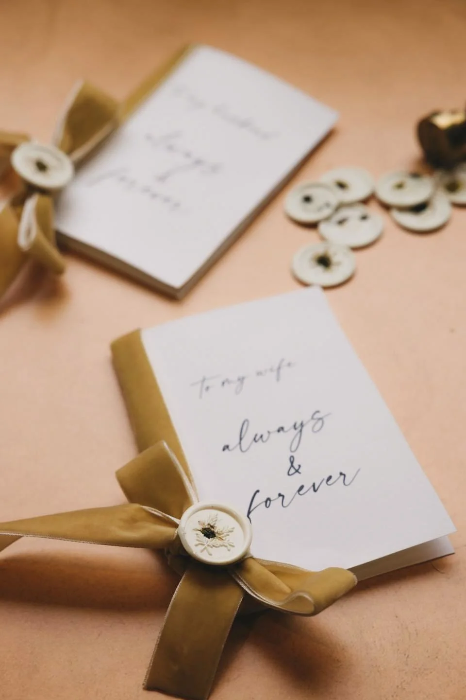 A close-up of a handwritten note on white paper with gold accents tied with a beige ribbon, surrounded by white buttons and a small jar in the background, on a pink surface.
