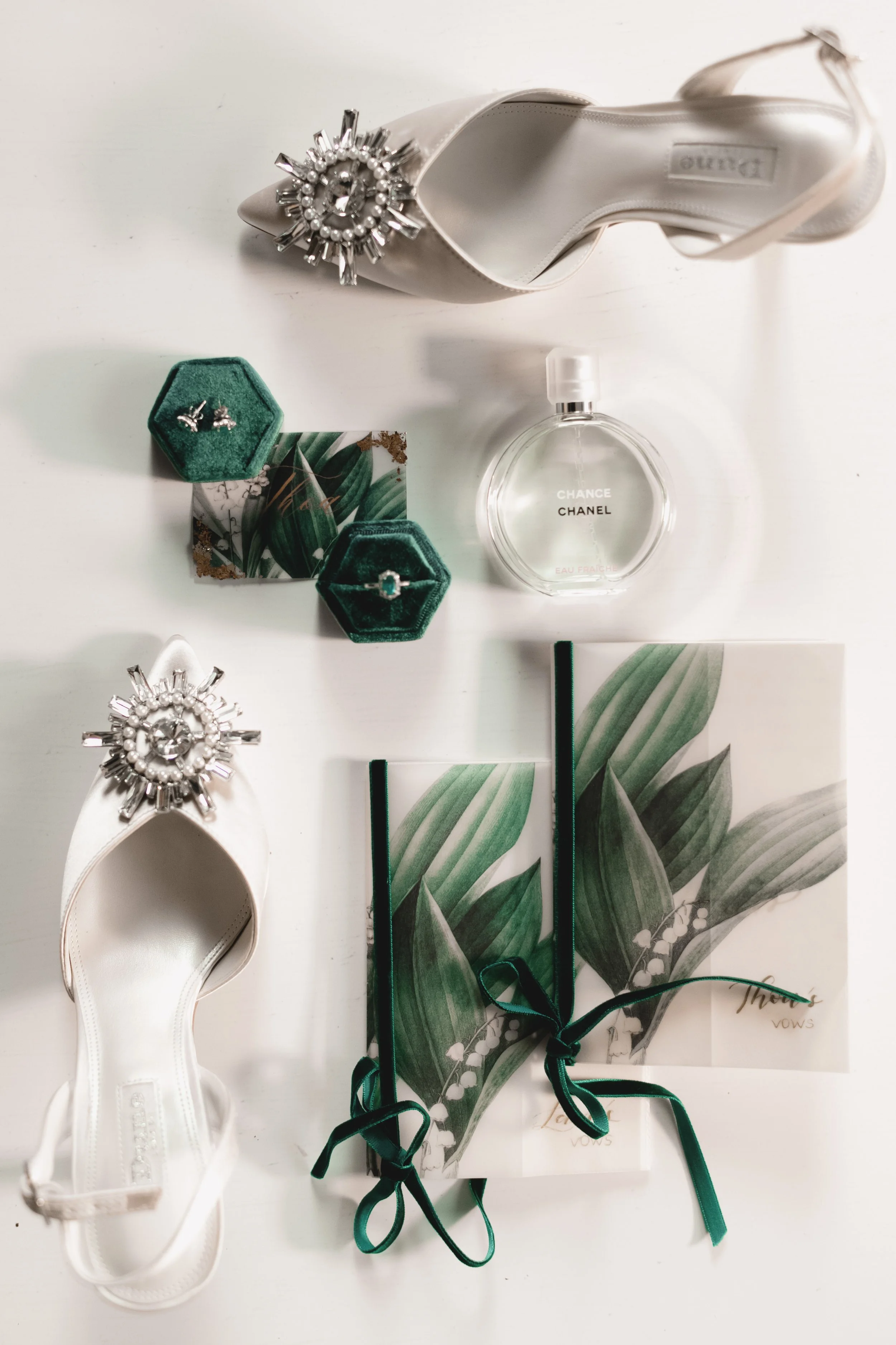 White high heel shoes with jeweled brooch accents, green velvet jewelry boxes with earrings and a ring, a bottle of Chanel Chance perfume, notebooks with green leaf designs and black ribbons, and a book titled 'There Vows' arranged on a white surface.