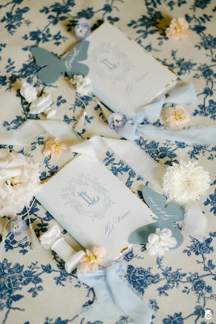 Wedding invitation suite decorated with flowers and butterflies on a blue and white patterned tablecloth.