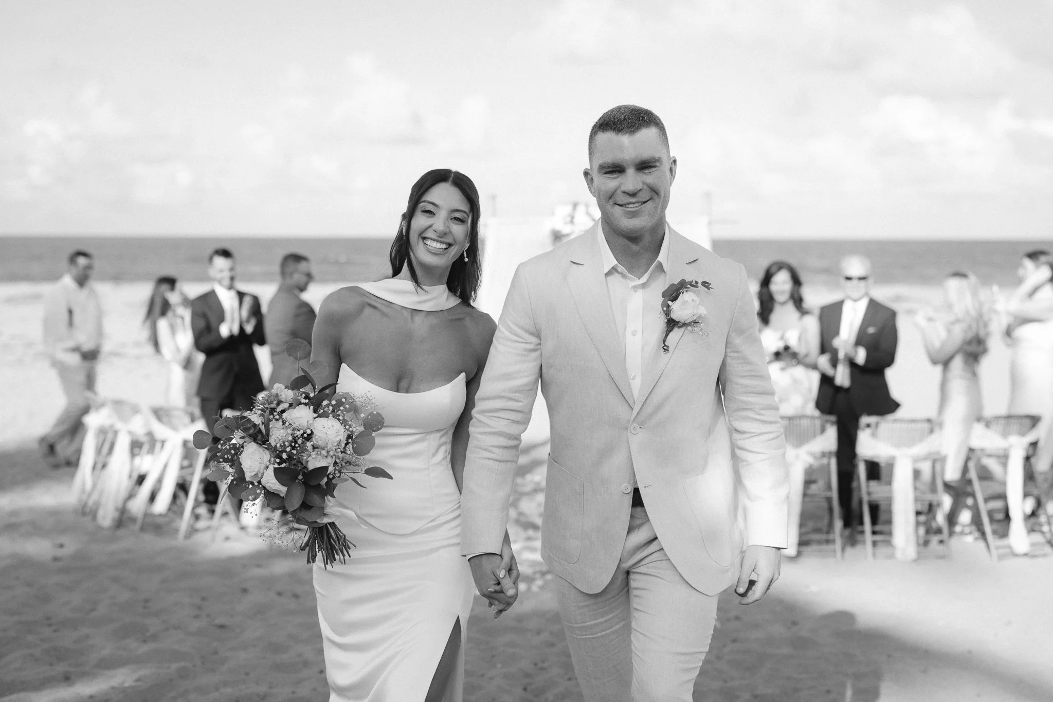 A newlywed couple walking hand-in-hand along the beach during their wedding ceremony, with wedding guests in the background.