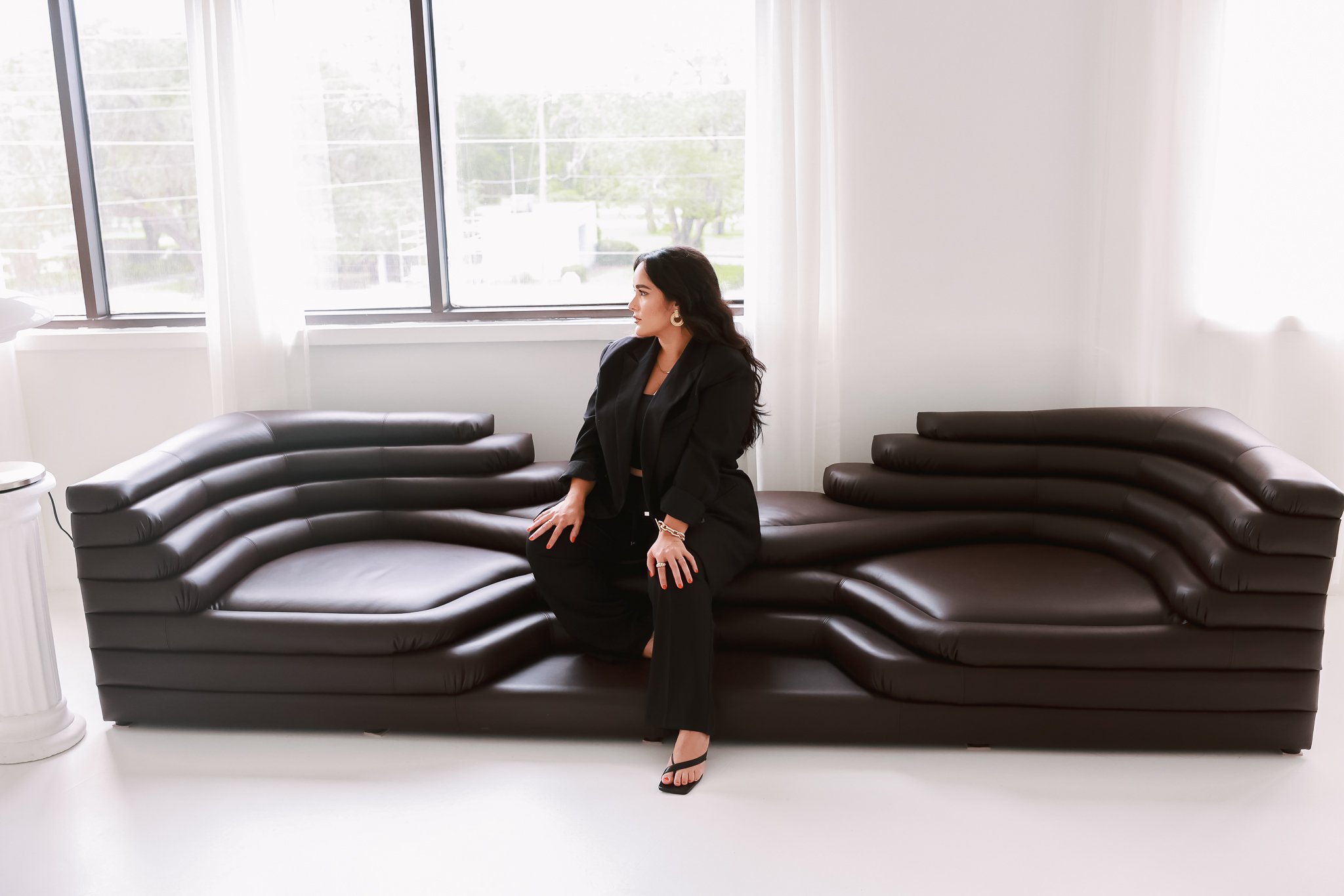 A woman with long dark hair, wearing a black blazer and pants, and high heels, sitting on a black leather sectional sofa in a bright room with large windows.