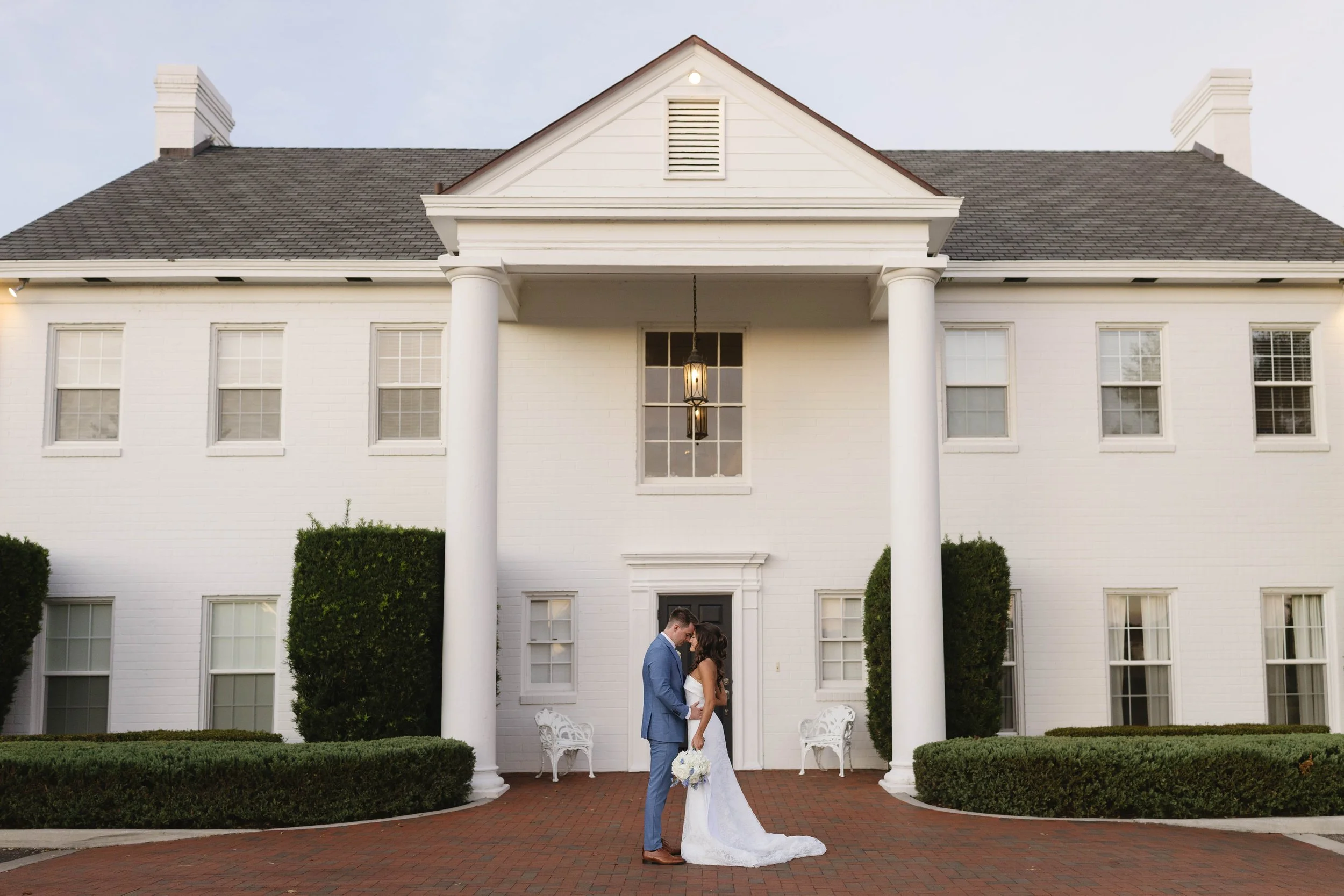 A bride and groom stand close together, holding hands, in front of a large white house with tall columns, multiple windows, and a porch light hanging between them. The bride is holding a bouquet and the groom is wearing a blue suit. The scene is outdoors on a brick patio, with neatly trimmed bushes.