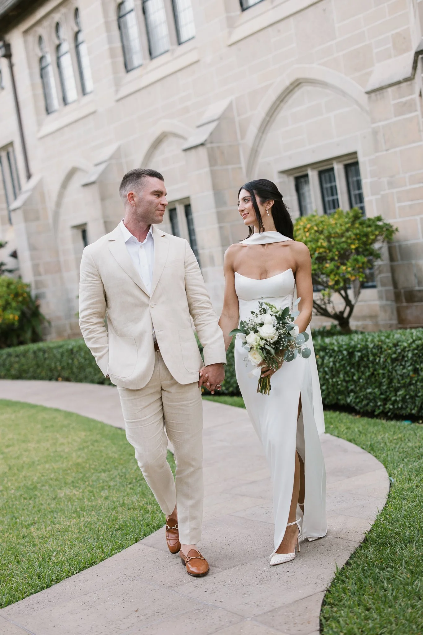 A married couple walking hand in hand outside a stone building, dressed in wedding attire, with the woman holding a bouquet of white flowers.