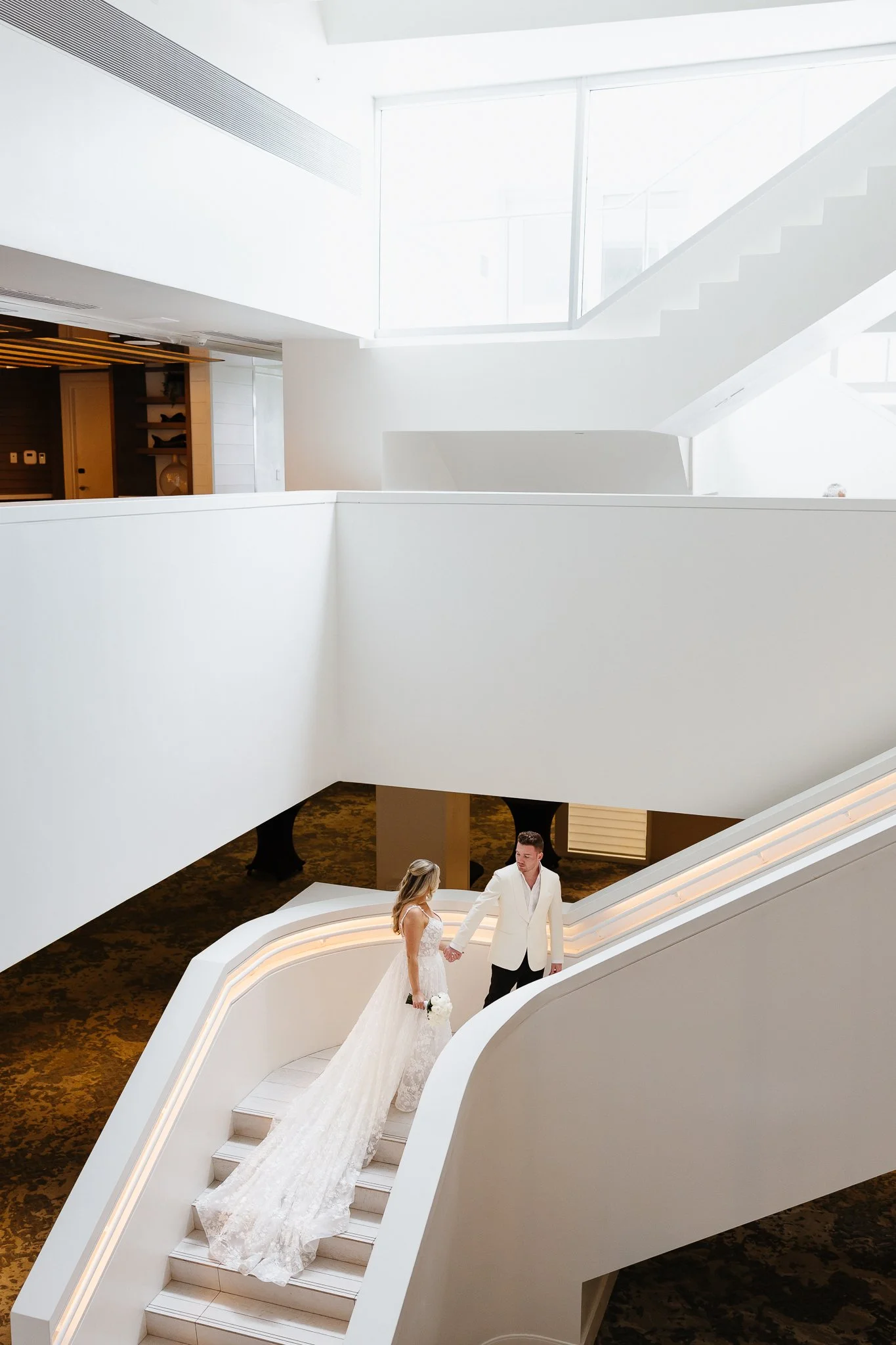 A bride and groom standing on white stairs inside a modern, bright venue with white walls and curved architecture, holding hands and looking at each other.