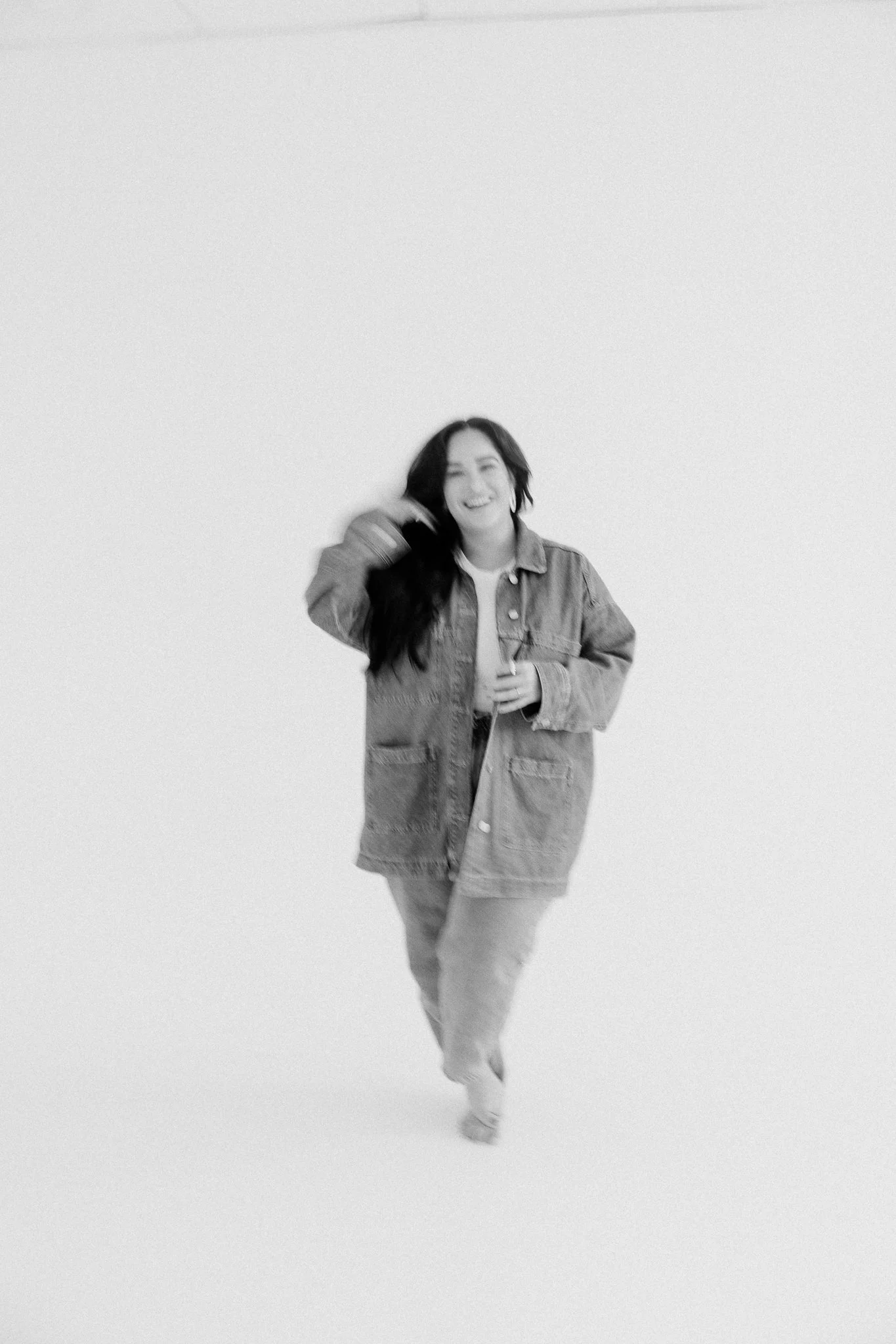 Black and white photo of a young woman with long dark hair, smiling and walking with one hand in her pocket and the other touching her hair, wearing a denim jacket and pants, against a plain light background.