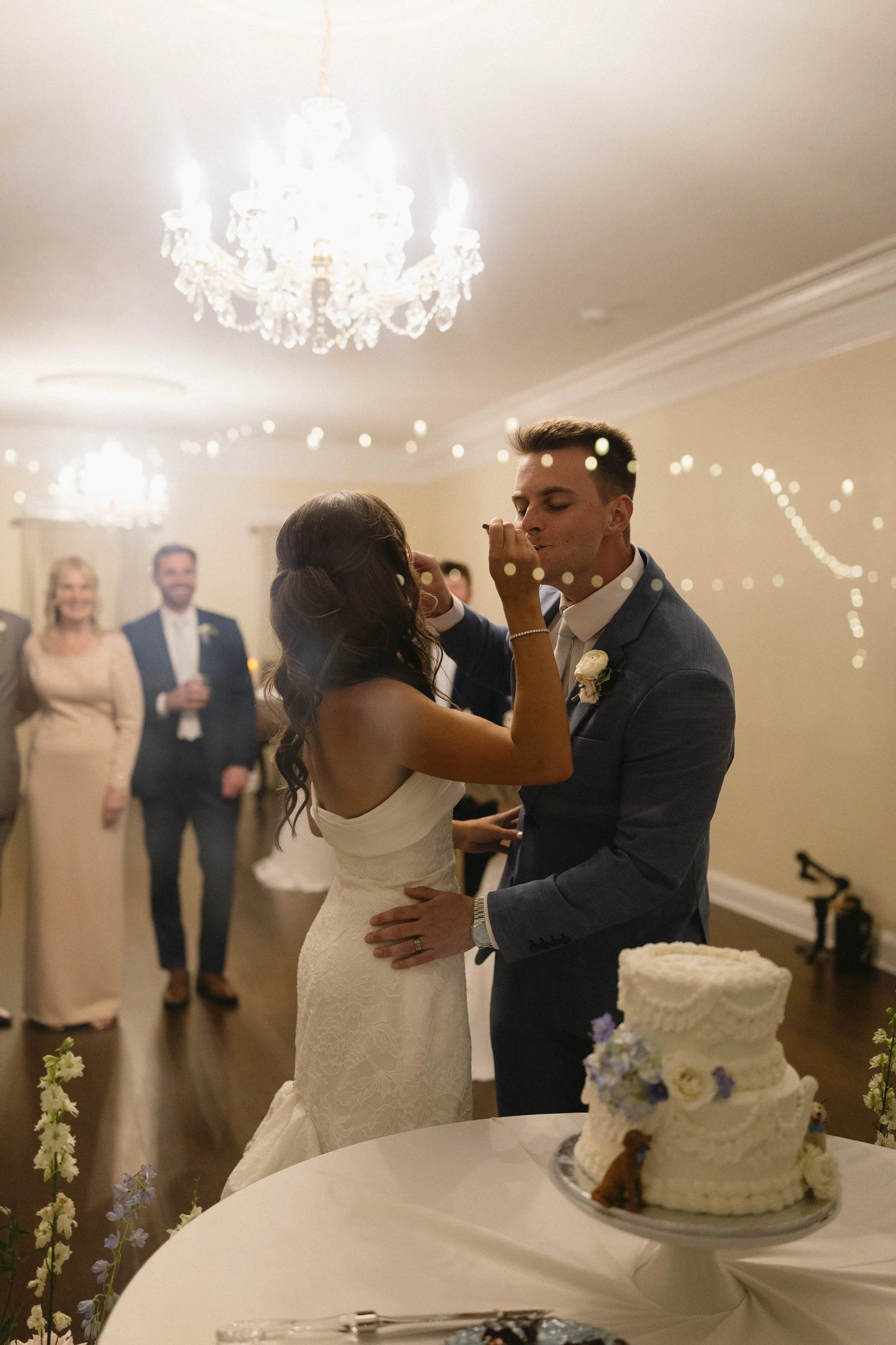 A bride and groom dancing at their wedding reception with guests in the background. A wedding cake with flowers is in the foreground.