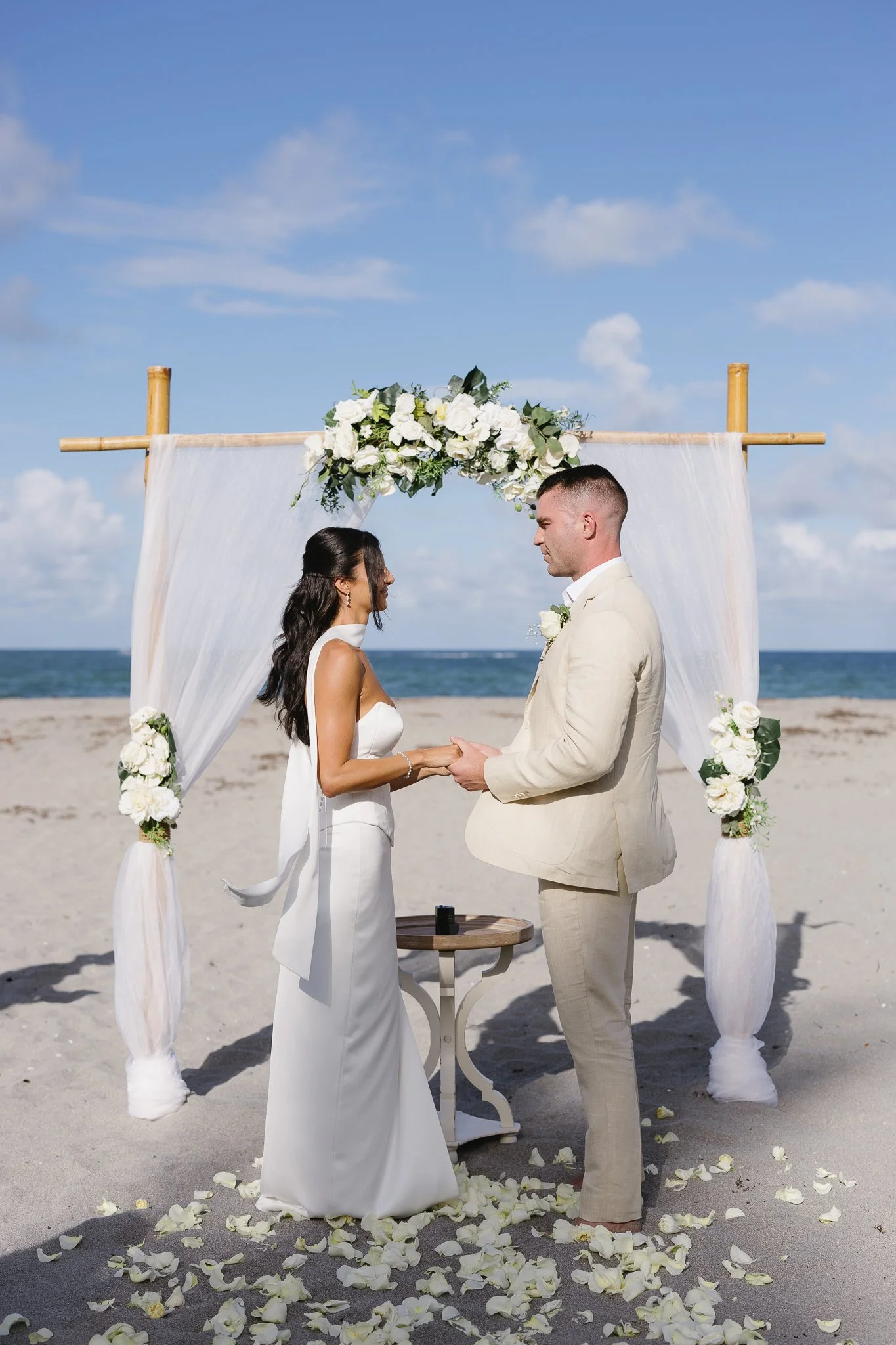A couple is standing face to face during a wedding ceremony on the beach, holding hands, under a wooden arch decorated with white flowers and draped fabric. Rose petals are scattered on the sand around them, with the ocean and sky in the background.