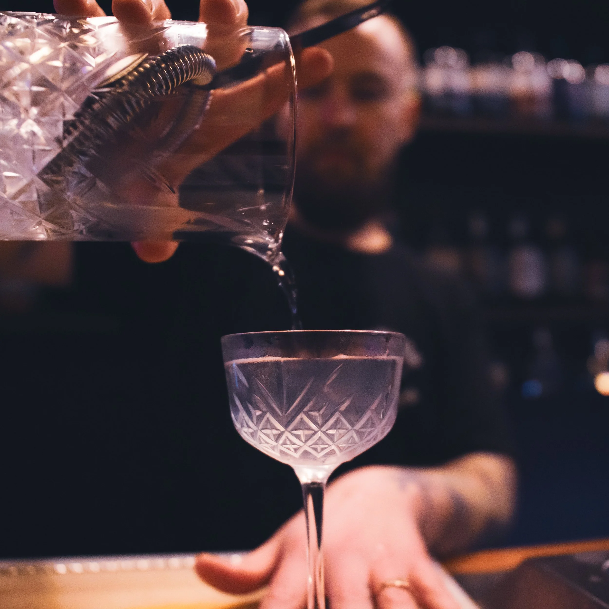 A person pours water from a crystal pitcher into a pink crystal cocktail glass in a dimly lit bar.