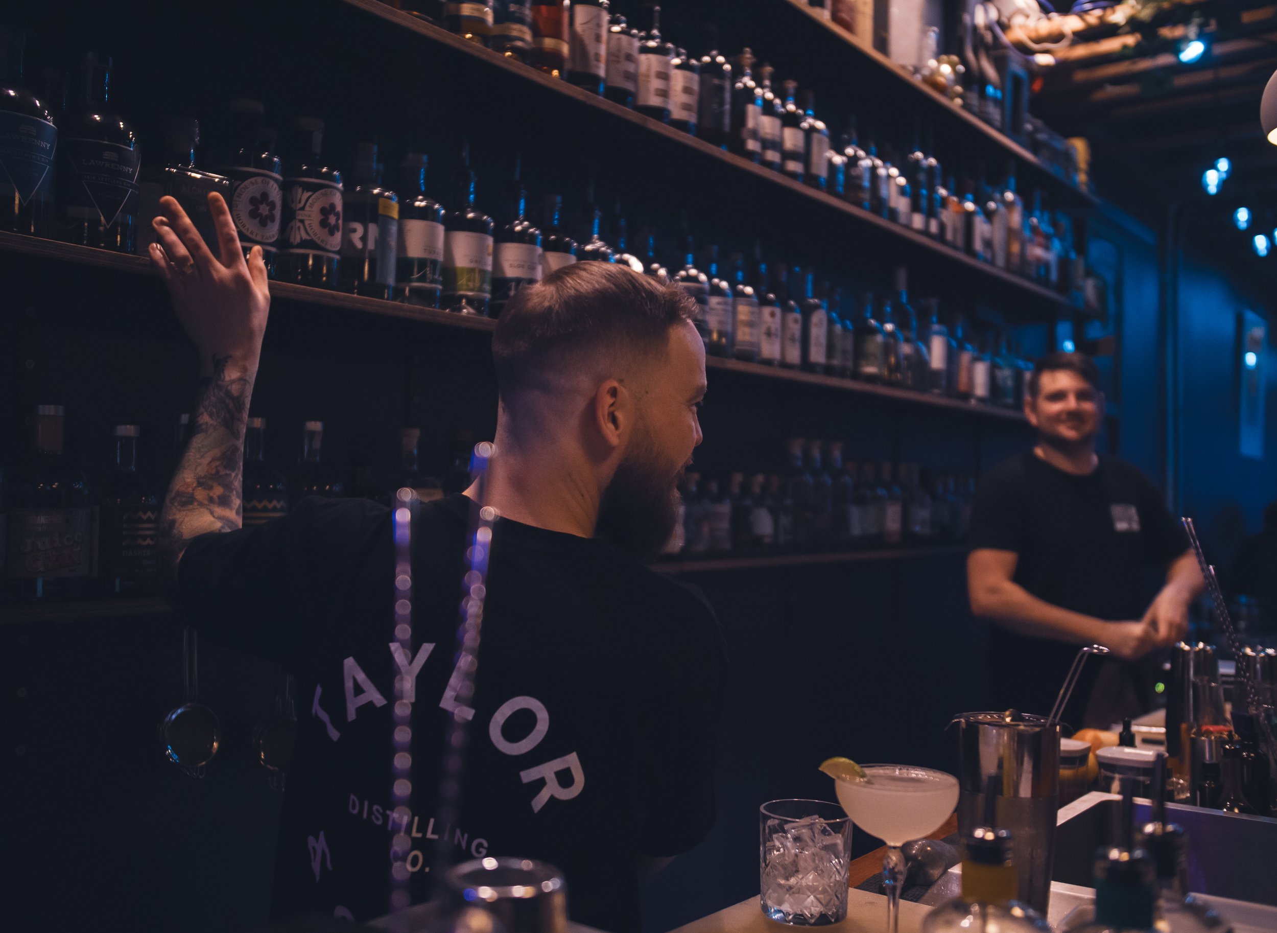Two bartenders behind a bar with shelves of liquor bottles, one with tattoos and a beard smiling, the other in the background preparing drinks, dimly lit with blue lighting.