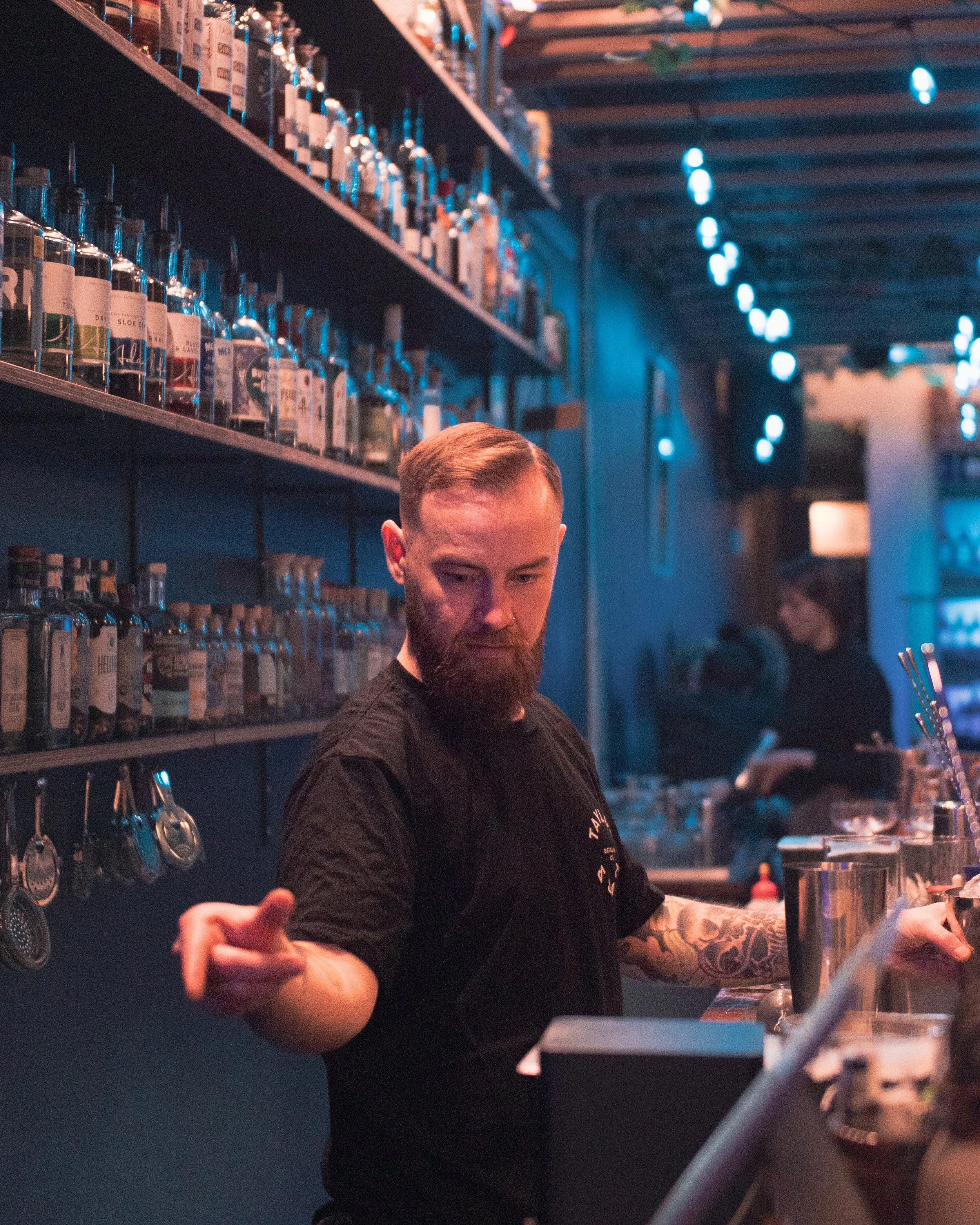 A bartender with a beard and tattooed arm working behind a bar with bottles on shelves in a dimly lit, blue-toned bar or club