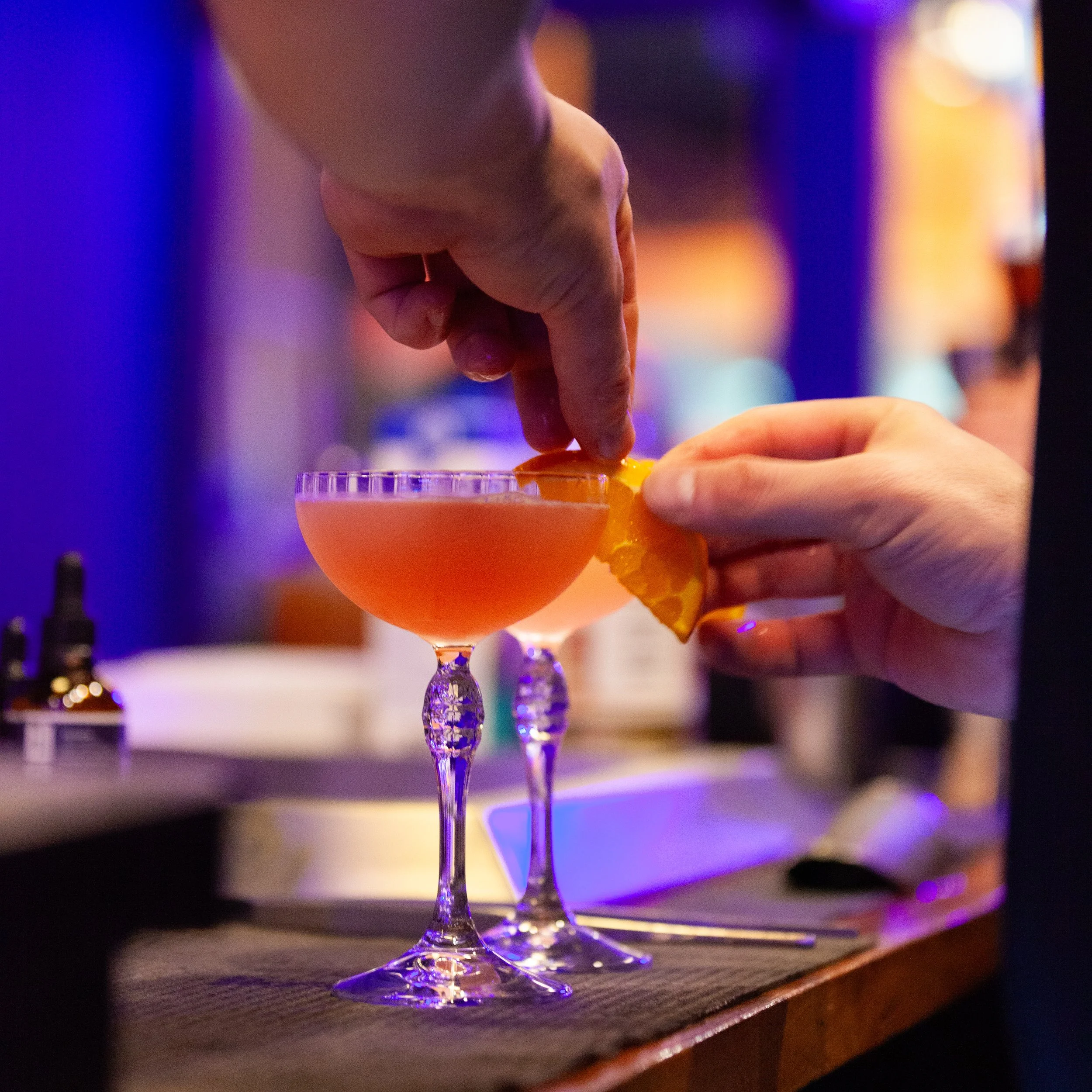 Two people preparing cocktails with orange garnish in a dimly lit bar setting.