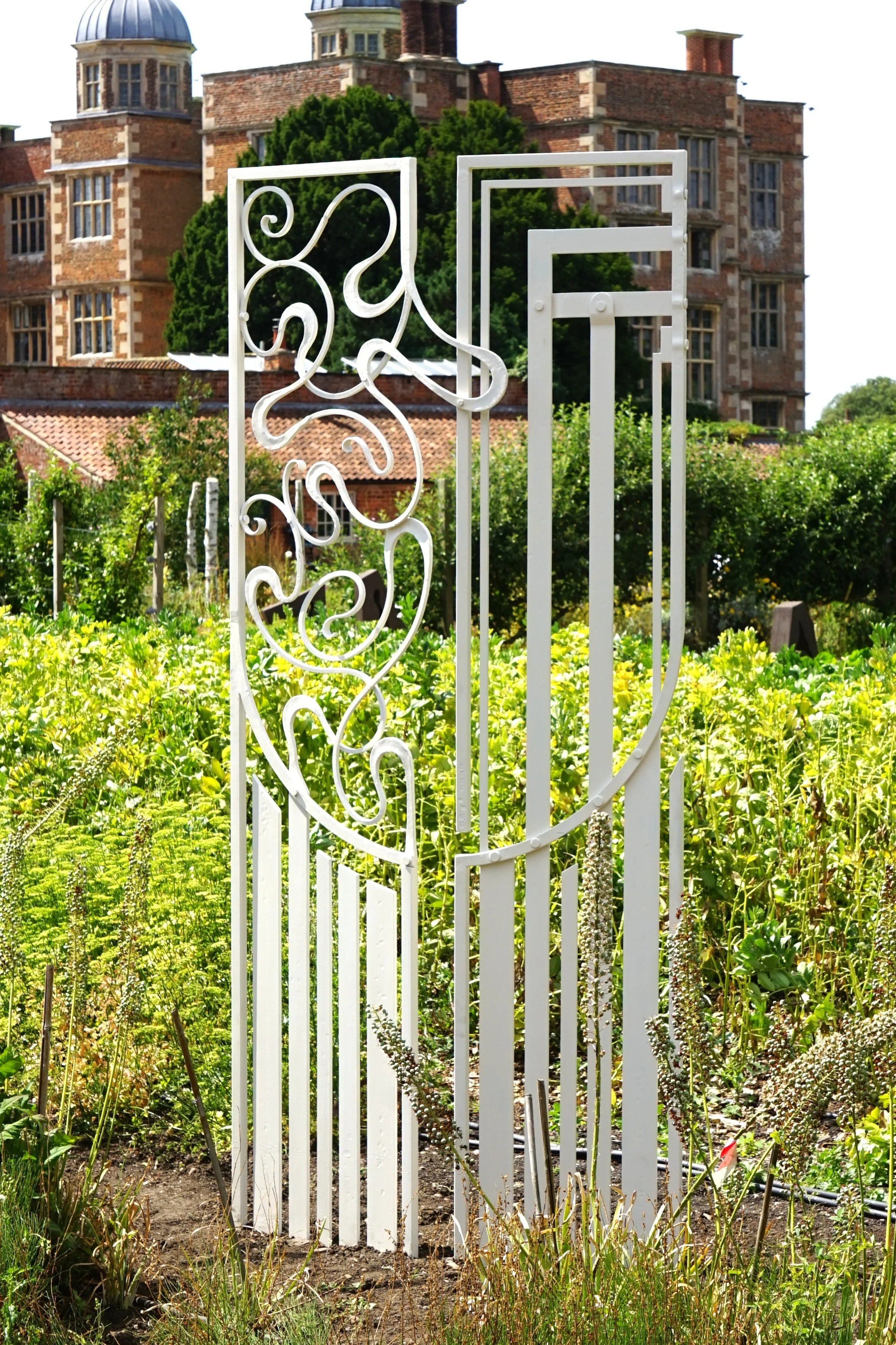 A white decorative garden gate with intricate swirl patterns stands open in a lush garden. In the background is a large brick mansion with tall windows and a domed tower.
