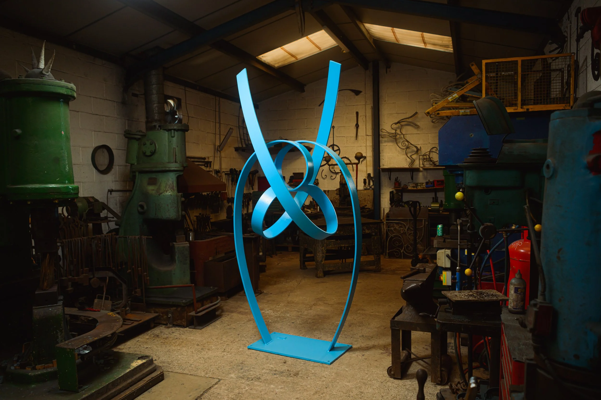 A large blue metal sculpture of intertwined loops and lines in an industrial workshop with tools, machinery, and storage shelves around. contemporary sculpture in a modern day forge