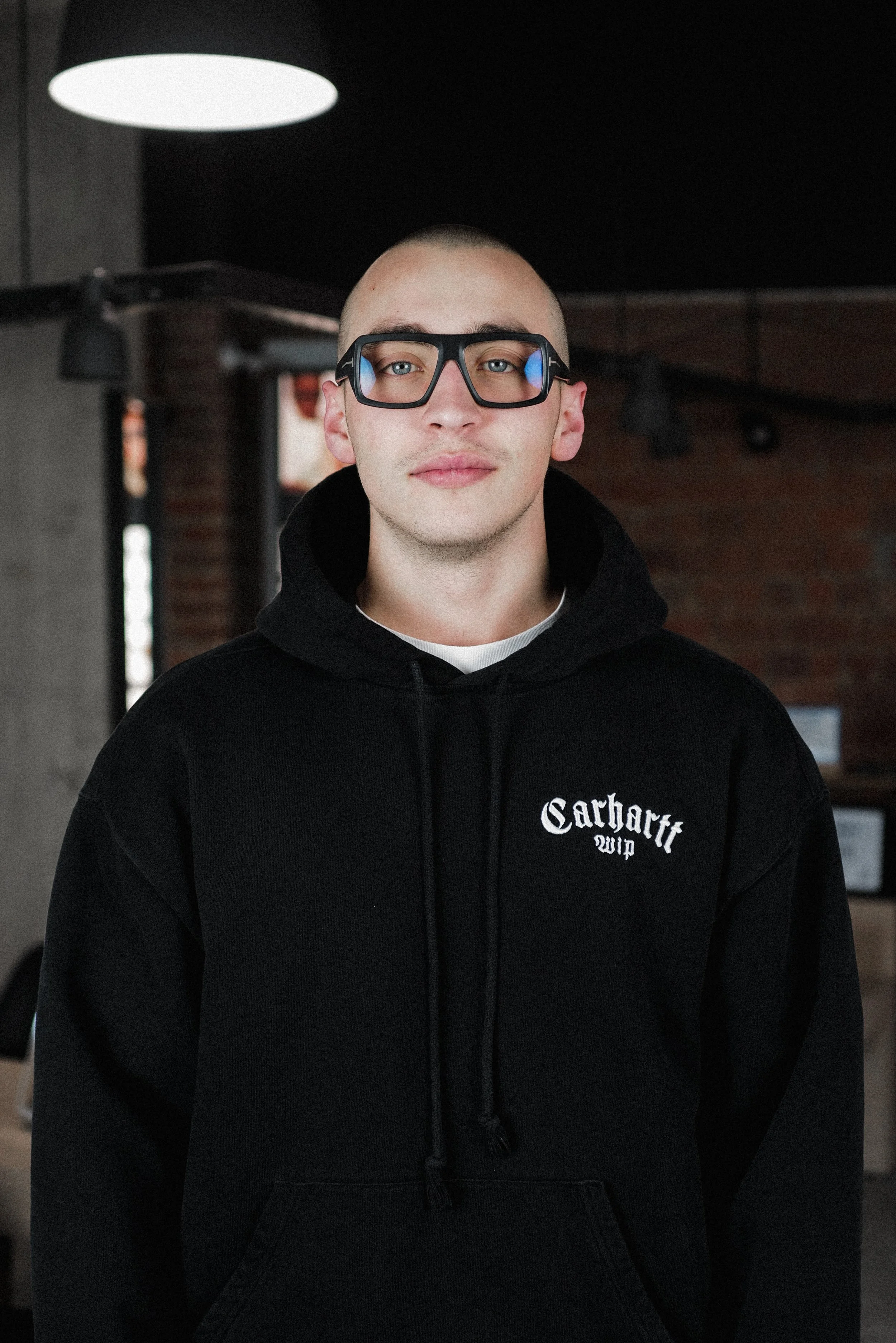 Bald man wearing black hoodie with "Carhartt WIP" logo and large black glasses, standing in a dimly lit room.