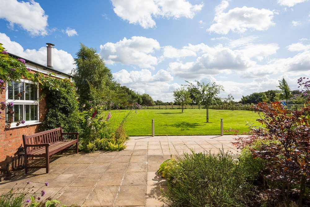  flagstone patio area with small planted up areas, beyond is a large fenced lawn area with two young trees in the centre  