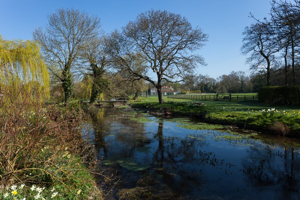  sunny day along a river bank with a house in the distance, fields on one side of the river and a grassy bank of the left, a small footbridge can be seen in the distance  