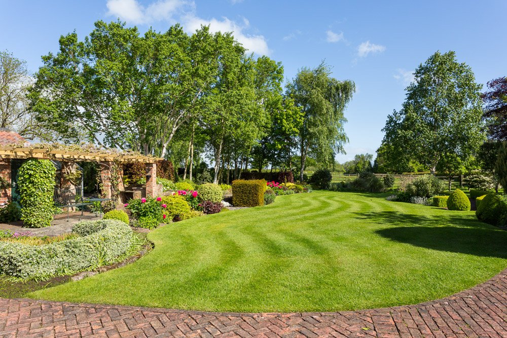  block paved foreground with neat lawn beyond, left hand side of garden neatly planted up with box hedges and small shrubs, tall trees boarder the left side 