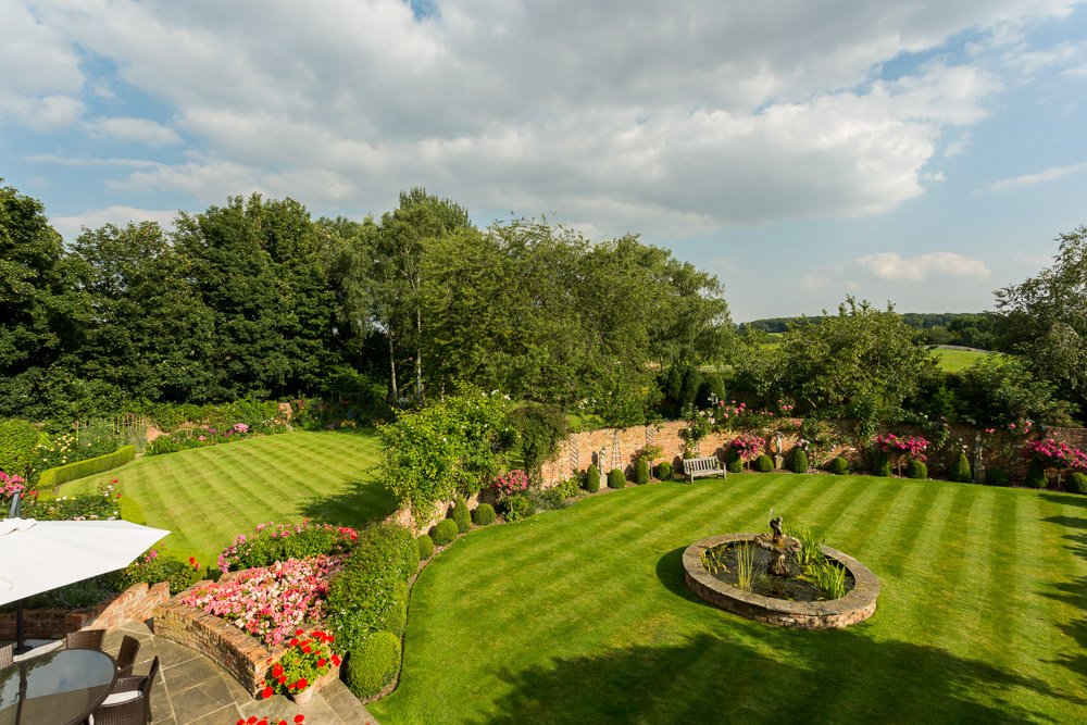  large walled garden with circular water feature and planted raised beds, beyond the wall is more law garden, tall trees and shrubs surround the whole garden 
