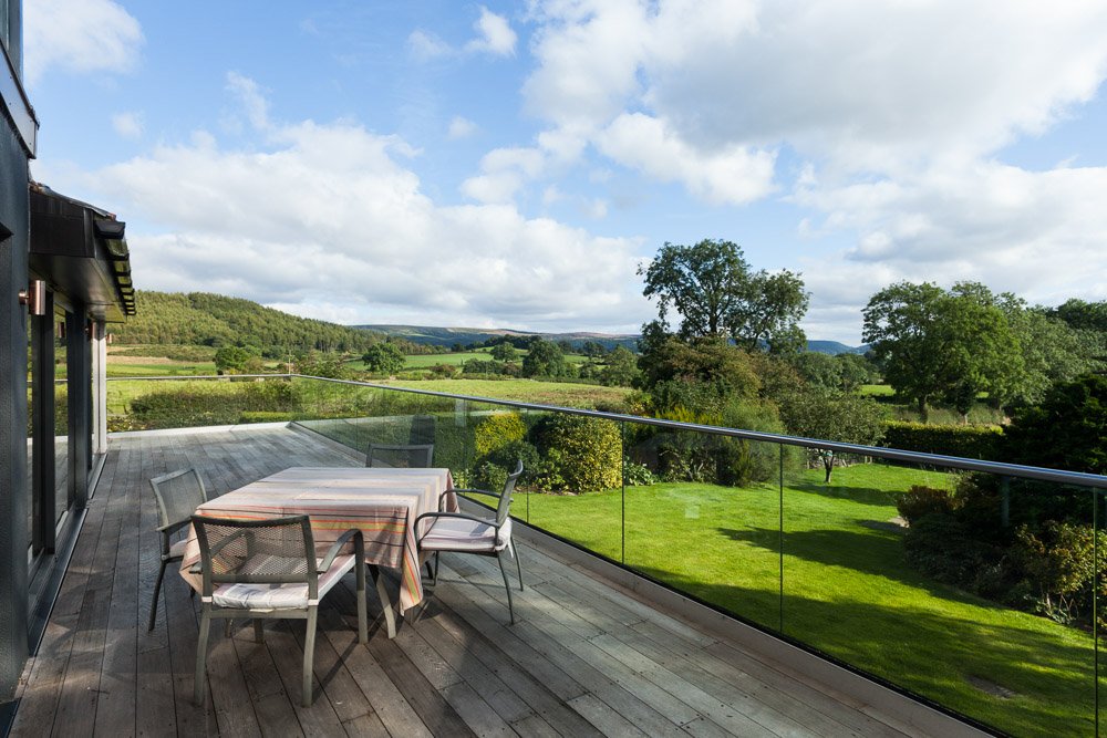 wooden balcony with glass barriers, small table and chairs, beyond the balcony rolling countryside for miles  