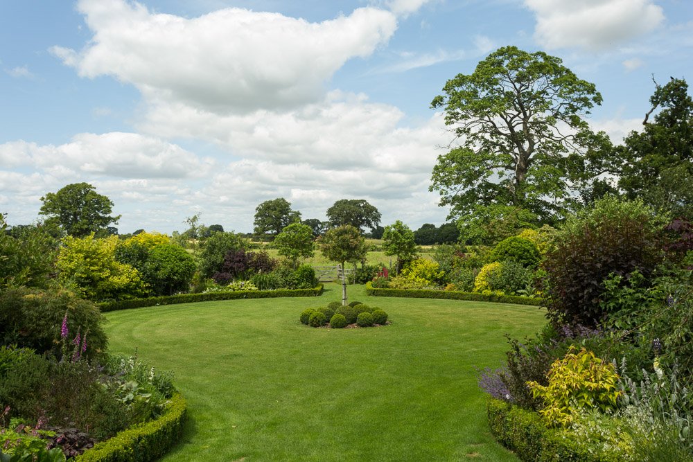  a circle patch of lawn with box hedging and planted shrubbery surrounding, small tree in the centre 