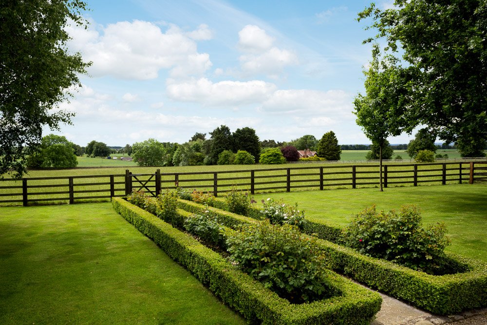  neat grass lawn with small box hedges surrounding planted up areas, beyond a wooden fence is miles of fields   