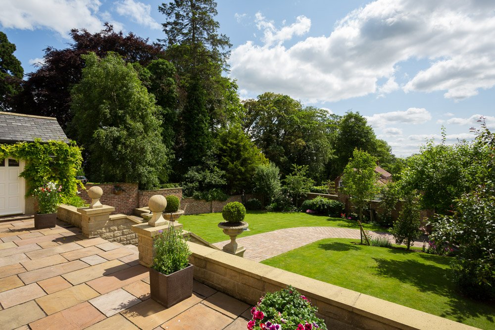  raised patio area above a neat lawn with block paving path through the middle, tall trees line the left hand side 