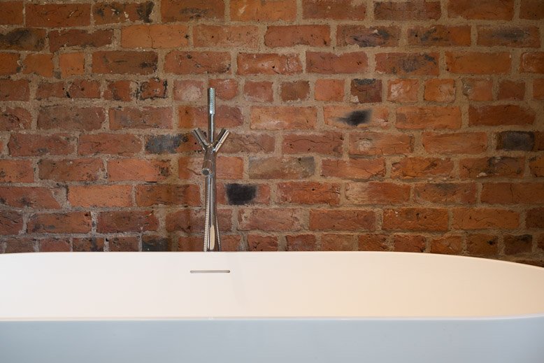  flat no contrast sleek modern silver tap over a curved white bathtub with bare brick wall behind 