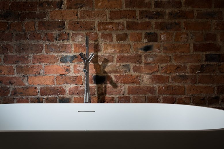  harshly lit high contrast sleek modern silver tap over a curved white bathtub with bare brick wall behind 