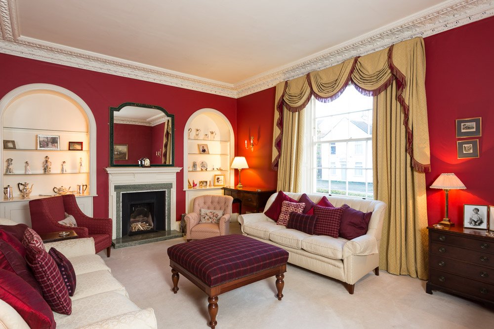  harshly lit  too bright drawing room with red walls, cream sofas and carpet, lit fireplace, gold draped curtains, purple chequered foot stool in centre of room 