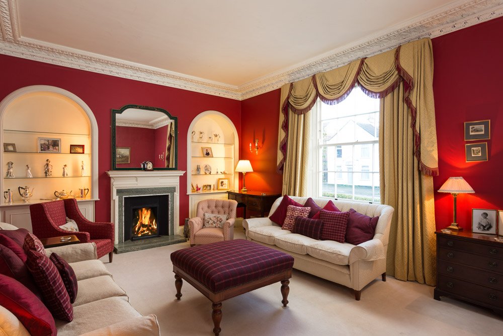  edited and well exposed drawing room with red walls, cream sofas and carpet, lit fireplace, gold draped curtains, purple chequered foot stool in centre of room 
