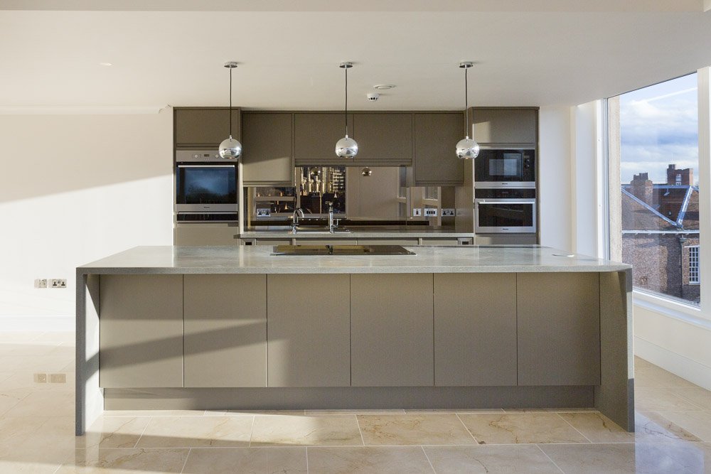  sleek modern apartment kitchen with grey cupboards and grey marble countertops, sun beams across the worksurfaces from large windows on the right  
