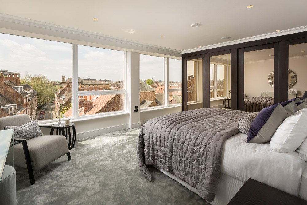  same modern apartment bedroom furnished with large bed covered in a grey blanket. grey arm chair sits below the panoramic windows overlooking York city centre, reflected in the mirrored wardrobes is a clock 