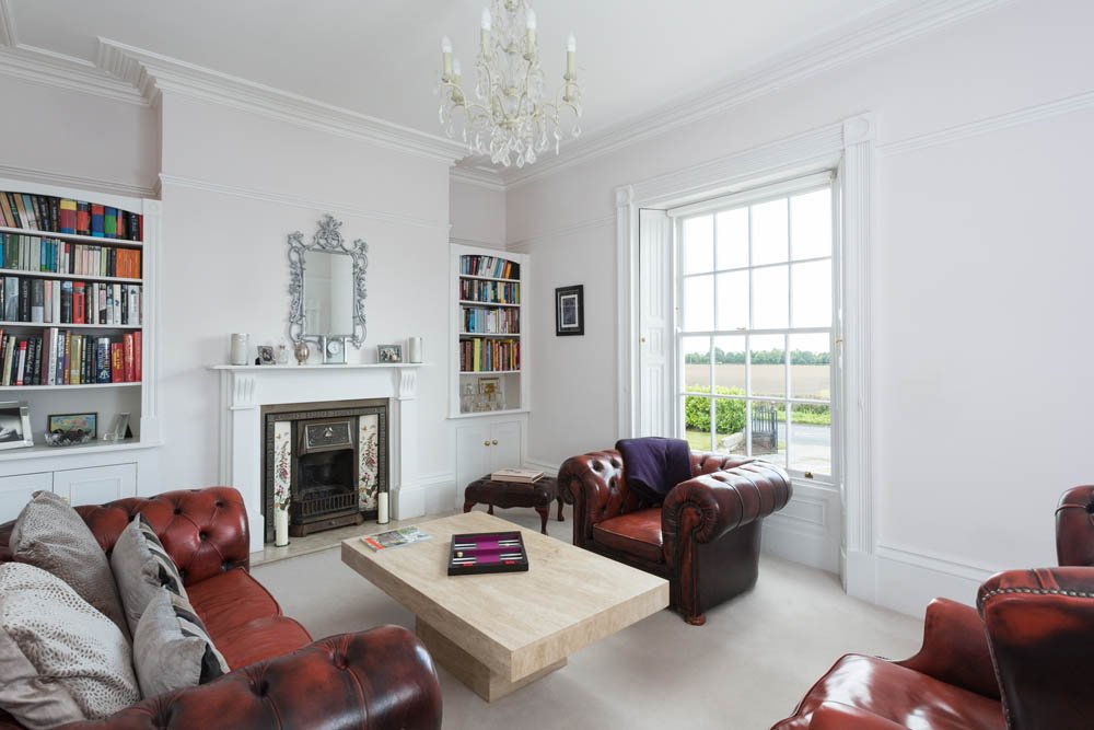  high ceiling white living room with dark red leather sofas, traditional fireplace and sash window 