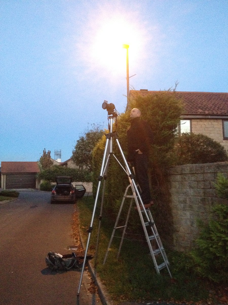  photographer on stepladder looking through camera on very tall tripod at dusk 