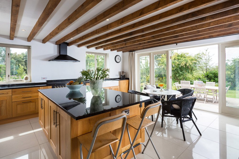  beamed ceiling kitchen with white tile flooring, wooden units, black countertops and small breakfast table, open doors lead to patio seating area 