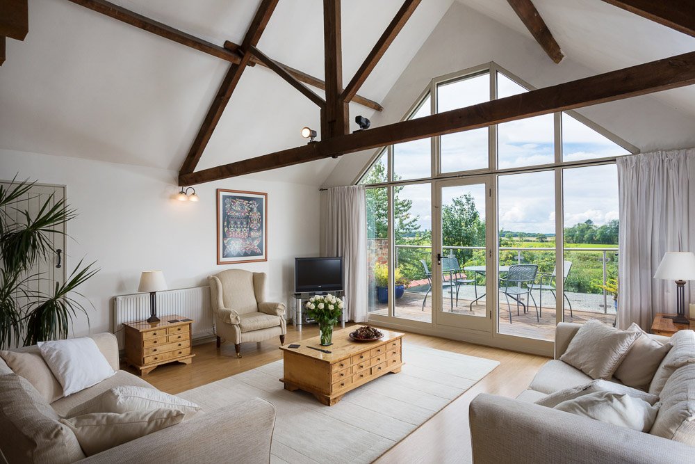  double height ceiling living room with wooden beams, wooden flooring, cream sofas, large glass wall looking over roof terrace and countryside  
