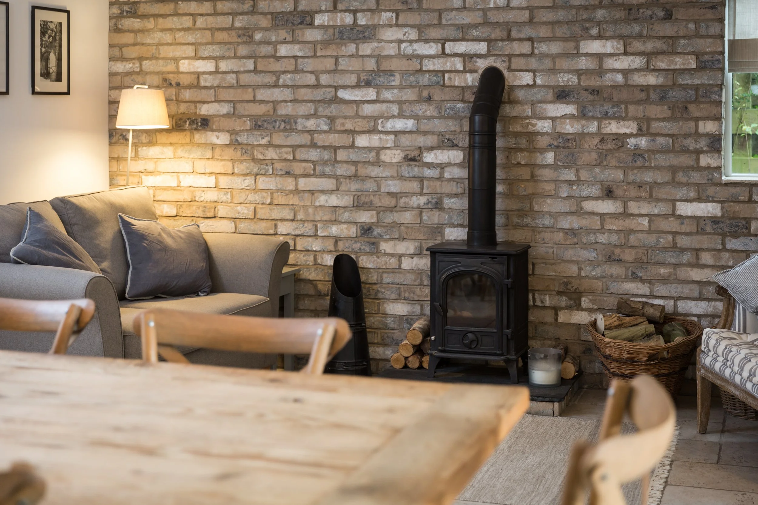 shallow depth of field image looking over dining table towards brick wall with log burner and cosy grey sofa